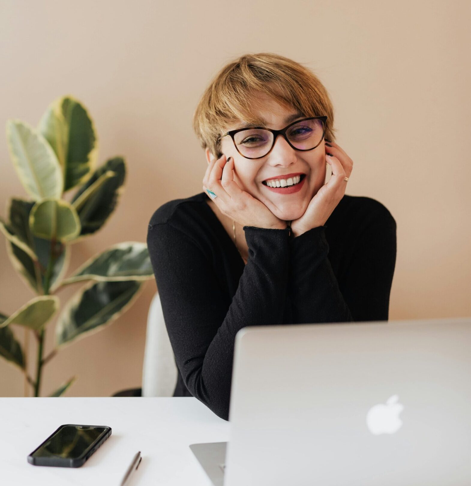 Cheerful woman in eyeglasses and black sweater smiling while working at a desk with a laptop indoors.
